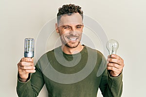 Young hispanic man holding lightbulb for inspiration and idea winking looking at the camera with sexy expression, cheerful and