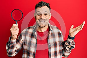 Young hispanic man holding magnifying glass looking for stain at clothes celebrating achievement with happy smile and winner