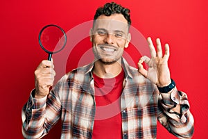 Young hispanic man holding magnifying glass looking for stain at clothes doing ok sign with fingers, smiling friendly gesturing