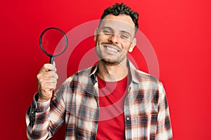 Young hispanic man holding magnifying glass looking for stain at clothes looking positive and happy standing and smiling with a