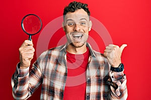 Young hispanic man holding magnifying glass looking for stain at clothes pointing thumb up to the side smiling happy with open