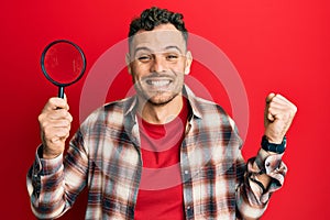 Young hispanic man holding magnifying glass looking for stain at clothes screaming proud, celebrating victory and success very