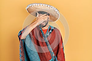 Young hispanic man holding mexican hat smiling and laughing with hand on face covering eyes for surprise