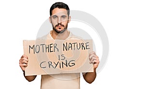 Young hispanic man holding mother nature is crying protest cardboard banner thinking attitude and sober expression looking self