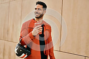 Young hispanic man holding motorcycle helmet and drinking coffee at the city