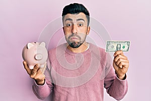 Young hispanic man holding one dollar banknote and piggy bank puffing cheeks with funny face