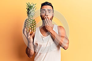 Young hispanic man holding pineapple covering mouth with hand, shocked and afraid for mistake