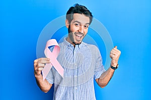 Young hispanic man holding pink cancer ribbon screaming proud, celebrating victory and success very excited with raised arms