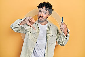 Young hispanic man holding pocket knife with angry face, negative sign showing dislike with thumbs down, rejection concept