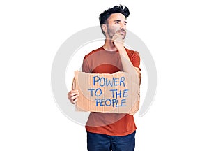 Young hispanic man holding power to the people banner serious face thinking about question with hand on chin, thoughtful about