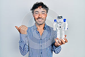 Young hispanic man holding robot toy pointing thumb up to the side smiling happy with open mouth