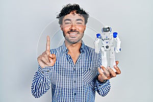 Young hispanic man holding robot toy smiling with an idea or question pointing finger with happy face, number one