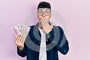 Young hispanic man holding saudi arabia riyal banknotes covering mouth with hand, shocked and afraid for mistake