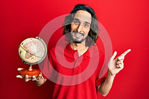 Young hispanic man holding small world ball smiling happy pointing with hand and finger to the side