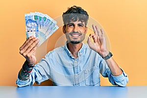 Young hispanic man holding south african rand banknotes sitting on the table doing ok sign with fingers, smiling friendly