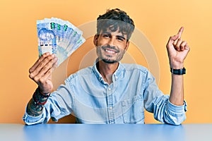 Young hispanic man holding south african rand banknotes sitting on the table smiling happy pointing with hand and finger to the