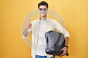 Young hispanic man holding suitcase going on summer vacation doing happy thumbs up gesture with hand