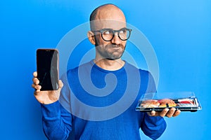 Young hispanic man holding take away food showing smartphone screen smiling looking to the side and staring away thinking