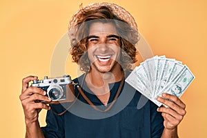 Young hispanic man holding vintage camera and dollars smiling and laughing hard out loud because funny crazy joke