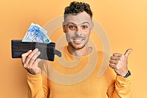 Young hispanic man holding wallet with south african rand banknotes pointing thumb up to the side smiling happy with open mouth