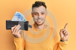 Young hispanic man holding wallet with south african rand banknotes smiling happy pointing with hand and finger to the side
