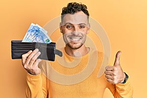 Young hispanic man holding wallet with south african rand banknotes smiling happy and positive, thumb up doing excellent and