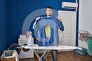 Young hispanic man ironing holding burned iron shirt at laundry room puffing cheeks with funny face