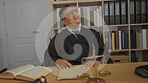 Young hispanic man in a judge\'s attire sitting in a courtroom office with a law book and scale on the desk photo