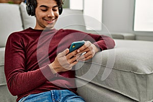 Young hispanic man listening to music sitting on the floor at home