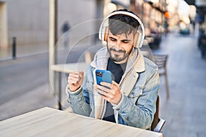 Young hispanic man listening to music sitting on table at coffee shop terrace