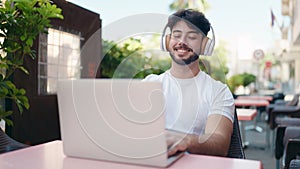 Young hispanic man listening to music sitting on table at coffee shop terrace