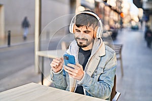 Young hispanic man listening to music sitting on table at coffee shop terrace