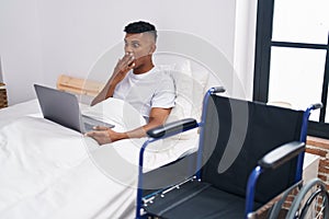 Young hispanic man lying on the bed, using wheelchair covering mouth with hand, shocked and afraid for mistake