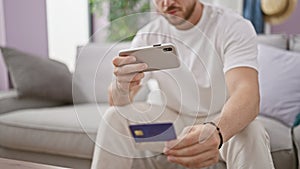 Young hispanic man make photo by smartphone to credit card sitting on sofa at home