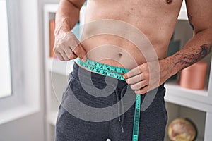 Young hispanic man measuring waist at bedroom