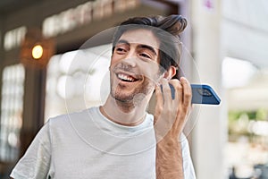 Young hispanic man miling confident listening audio message by the smartphone at street