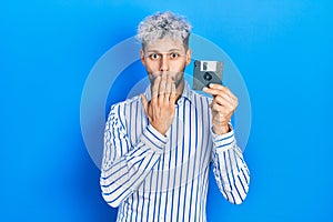 Young hispanic man with modern dyed hair holding floppy disk covering mouth with hand, shocked and afraid for mistake