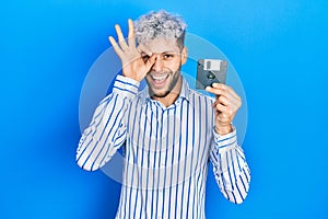 Young hispanic man with modern dyed hair holding floppy disk smiling happy doing ok sign with hand on eye looking through fingers