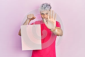 Young hispanic man with modern dyed hair holding shopping bag with open hand doing stop sign with serious and confident