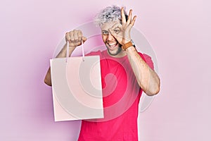 Young hispanic man with modern dyed hair holding shopping bag smiling happy doing ok sign with hand on eye looking through fingers