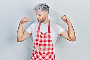 Young hispanic man with modern dyed hair wearing apron showing arms muscles smiling proud