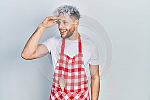 Young hispanic man with modern dyed hair wearing apron very happy and smiling looking far away with hand over head
