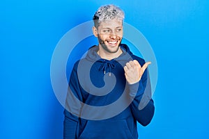 Young hispanic man with modern dyed hair wearing casual blue sweatshirt pointing thumb up to the side smiling happy with open