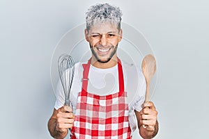Young hispanic man with modern dyed hair wearing cook apron holding baker whisk and spoon winking looking at the camera with sexy