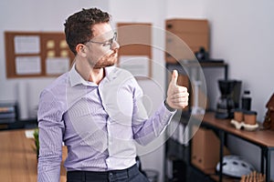 Young hispanic man at the office looking proud, smiling doing thumbs up gesture to the side