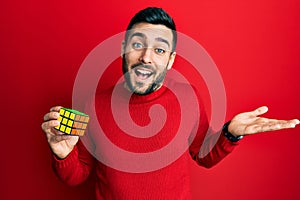 Young hispanic man playing colorful puzzle cube intelligence game celebrating achievement with happy smile and winner expression