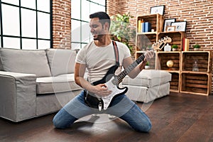 Young hispanic man playing electrical guitar with knees on floor at home