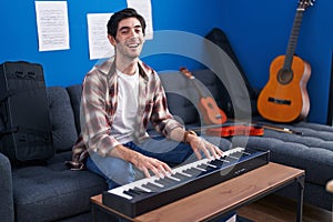 Young hispanic man playing piano at music studio smiling and laughing hard out loud because funny crazy joke