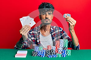 Young hispanic man playing poker holding cards and casino chips relaxed with serious expression on face