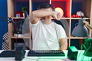 Young hispanic man playing video games covering eyes with arm, looking serious and sad